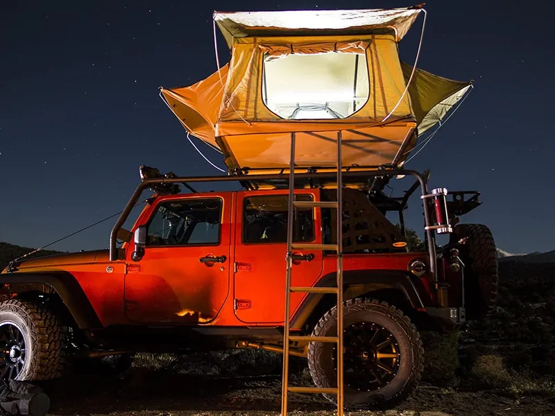 Smittybilt Overlander softshell fold-out rooftop tent open and lit from inside above an orange Jeep Wrangler under a starry night sky